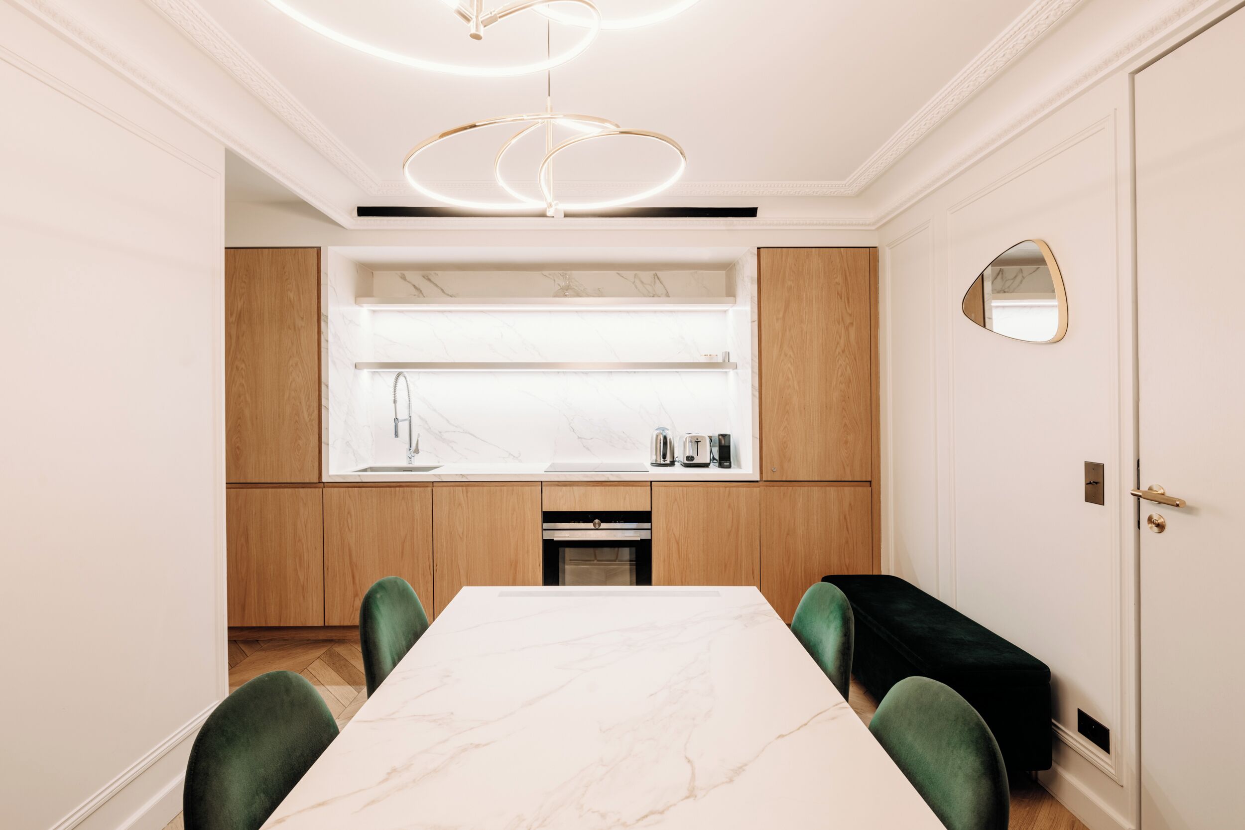 A modern dining area with a marble table, green upholstered chairs, wooden cabinets, and a built-in kitchen with a sink and countertop appliances.