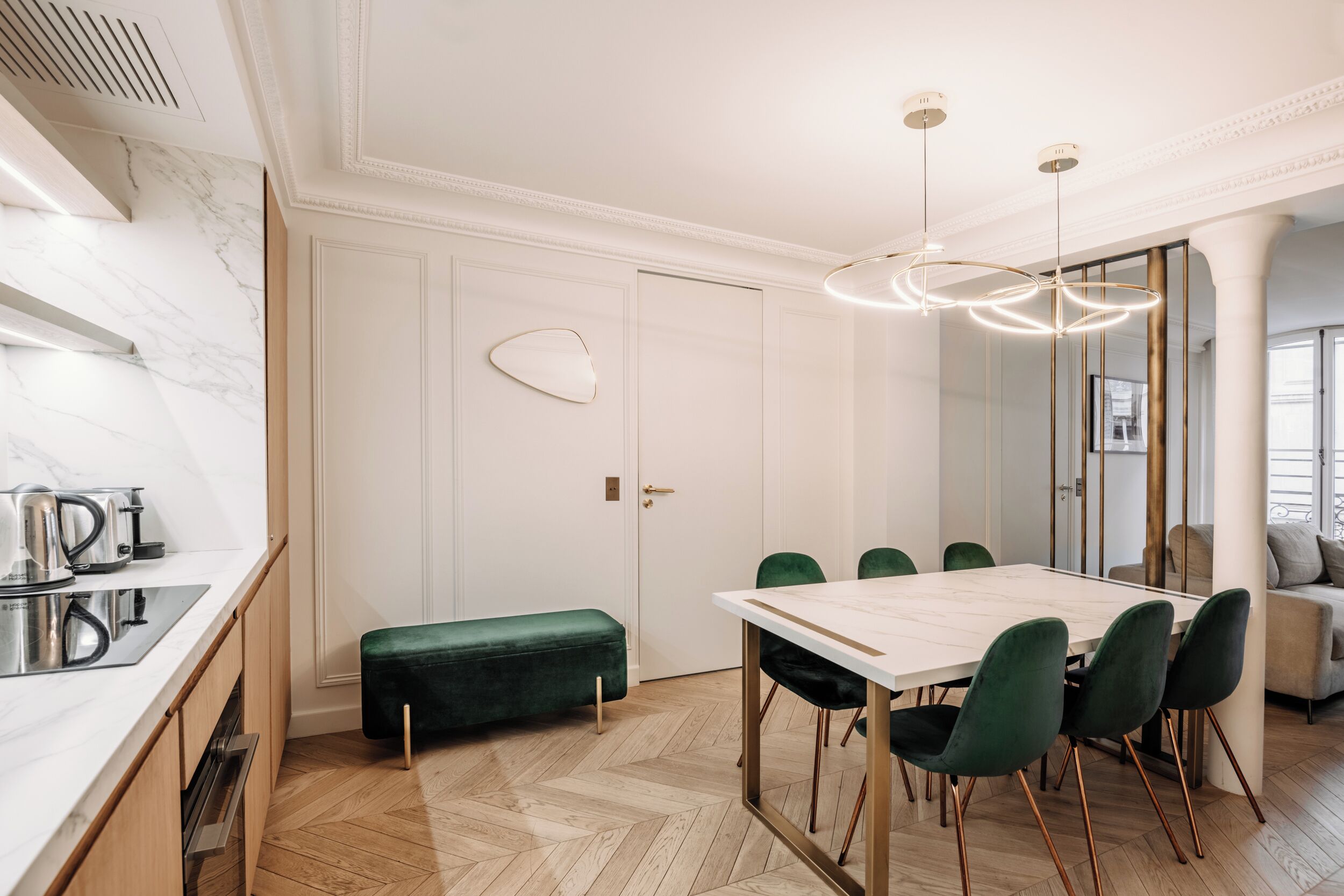 Un comedor moderno con encimera de mármol, suelo de madera en espiga, banca tapizada de verde y una mesa de comedor blanca con sillas verdes.