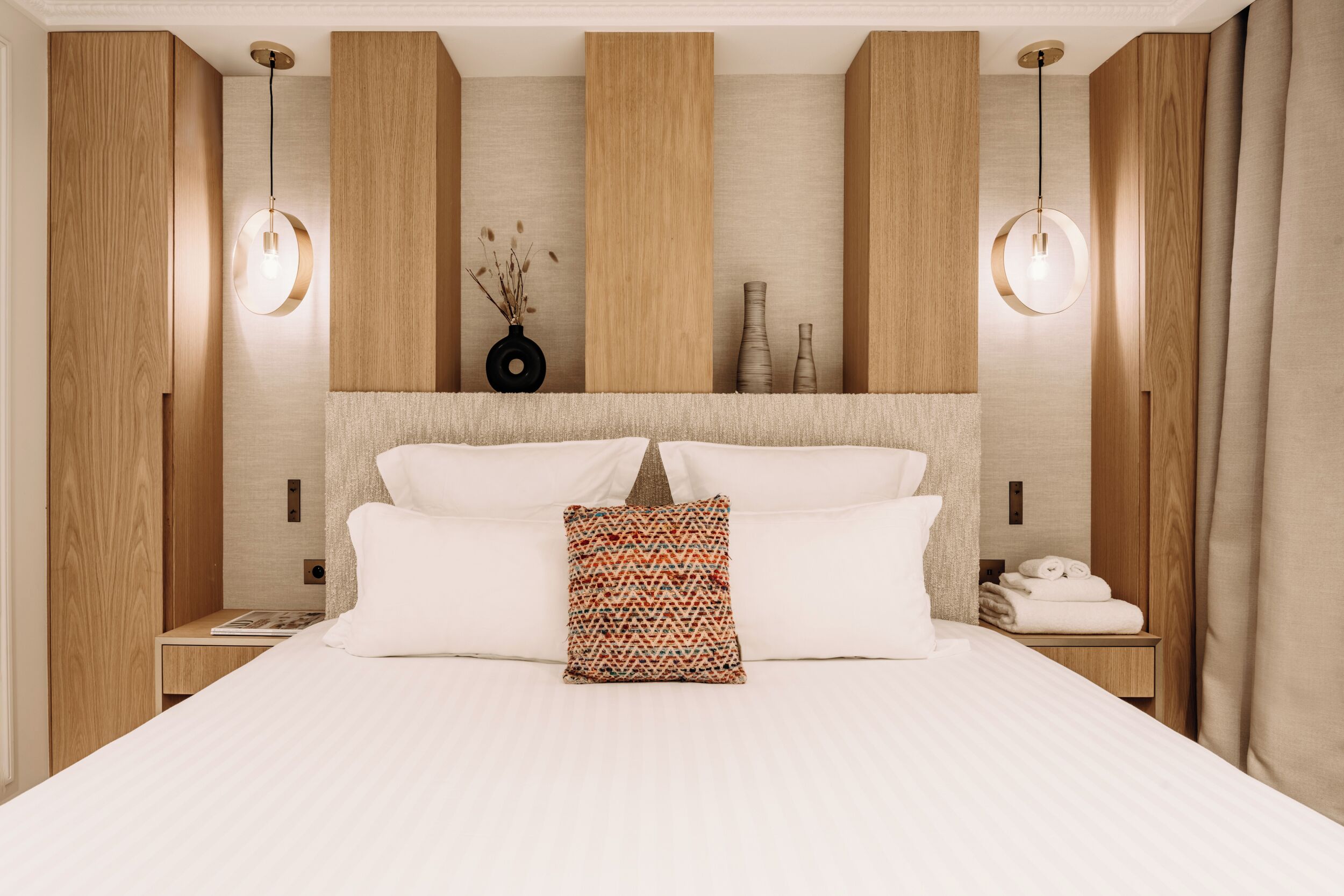 Una habitación de hotel moderna con una cama grande, paneles de madera, dos lámparas colgantes, un jarrón decorativo y toallas ordenadamente dobladas sobre una mesita de noche.