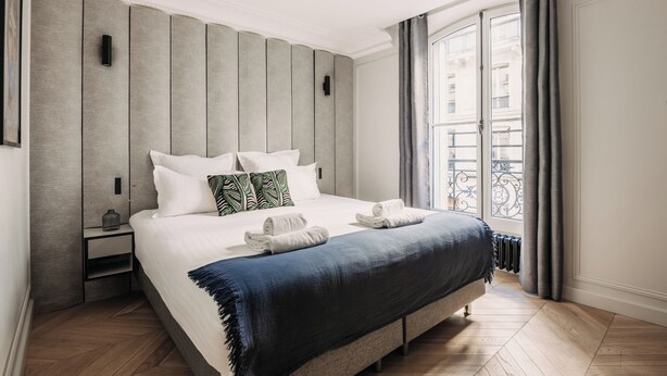 A modern hotel room with a large bed, a nightstand, a radiator, and a view of a balcony with a railing. - HIGHSTAY - Luxury Serviced Apartments - Louvre-Rivoli (Paris)