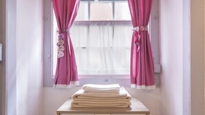 A room with a window, pink curtains, and a stack of white towels on a wooden table.