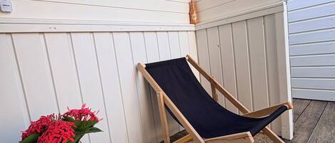 Une terrasse en bois avec un seul fauteuil de relaxation noir, une petite table sur laquelle est posé un vase de fleurs rouges, et un panneau accroché au mur.