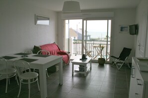 Ein Essbereich mit einem weißen Tisch und Stühlen, einem kleinen Couchtisch und einer Couch mit roter Decke. Ein Balkon bietet Blick auf das Meer.