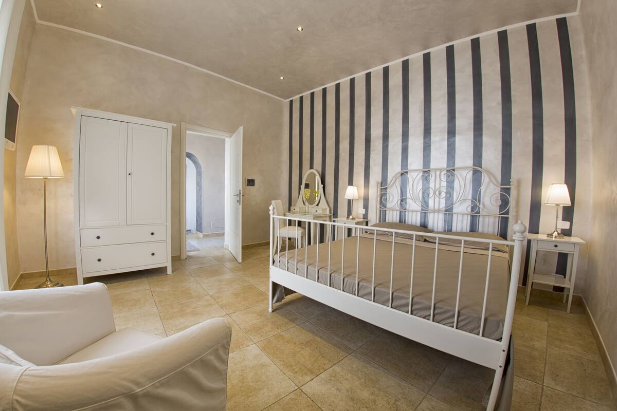 A bedroom with a white bed, a white chair, a bedside table with a lamp, a wardrobe, and a striped wall.