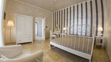 A bedroom with a white bed, a white chair, a bedside table with a lamp, a wardrobe, and a striped wall.