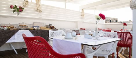 An outdoor dining area with white tables and red chairs, a bar area with food and drinks, and a view of buildings in the background.