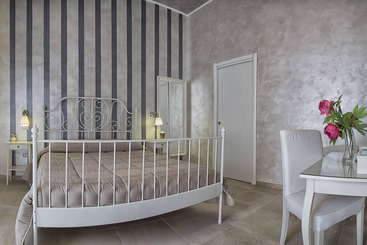A bedroom with a white metal bed, a bedside table with a lamp, a mirror, and a vase with flowers on a table.