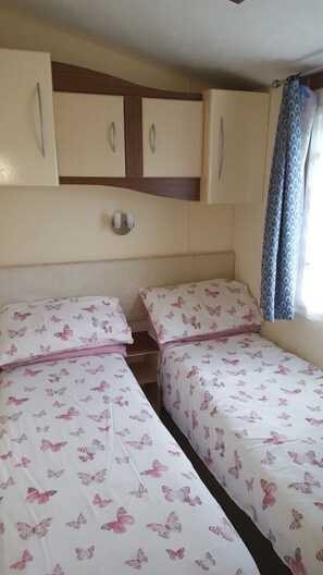 A bedroom with two beds, each with butterfly-patterned bedding, a wooden headboard, and a small bedside table with a lamp. - Welcome to my Caravan (Bude)