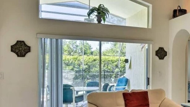 A living room with a high ceiling, a beige sofa, and a large sliding glass door leading to an outdoor patio. - Achieve your Magical Vacation! 12 Mins to Disney! (Kissimmee)