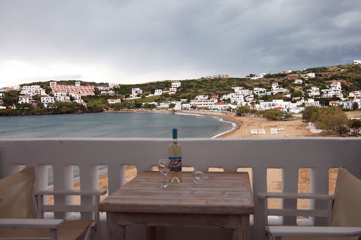 A terrace with a table, a bottle, and a glass. The terrace overlooks a beach and a town with white buildings.