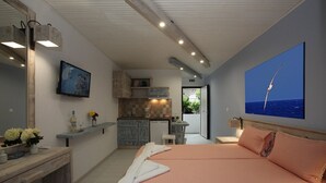 A bedroom with a bed, a desk, a chair, a mirror, a TV, a kitchen area, and a view of the sea. - Bella Vista Design Studios (Andros)