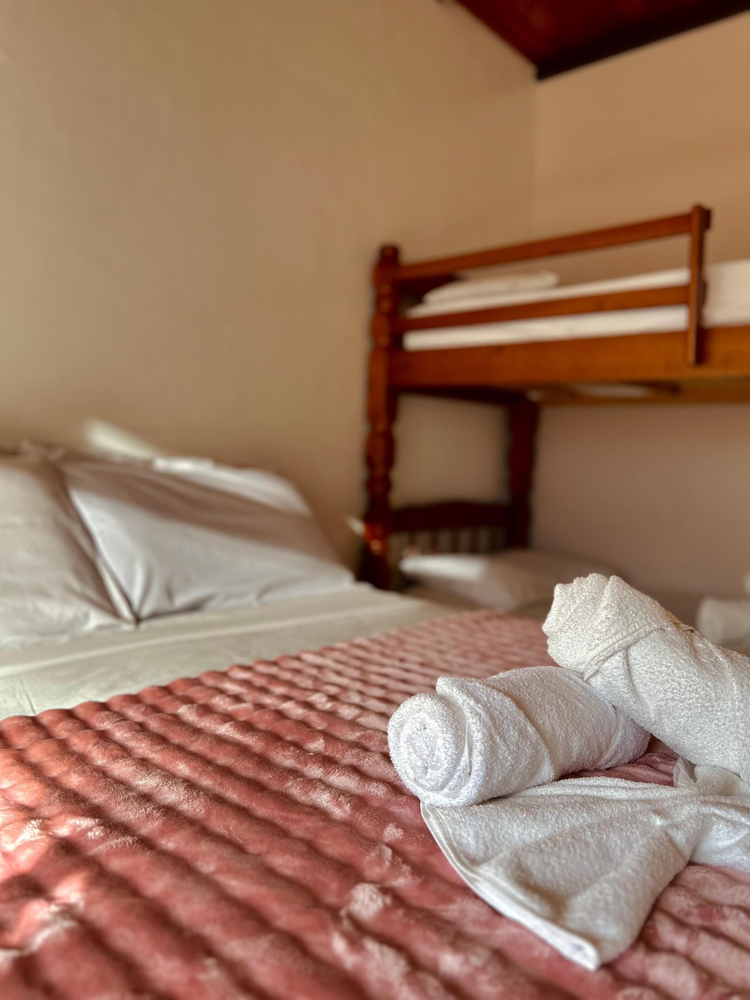 A hotel room with a bunk bed, a neatly made bed with white linens, and a rolled-up towel on the bed.
