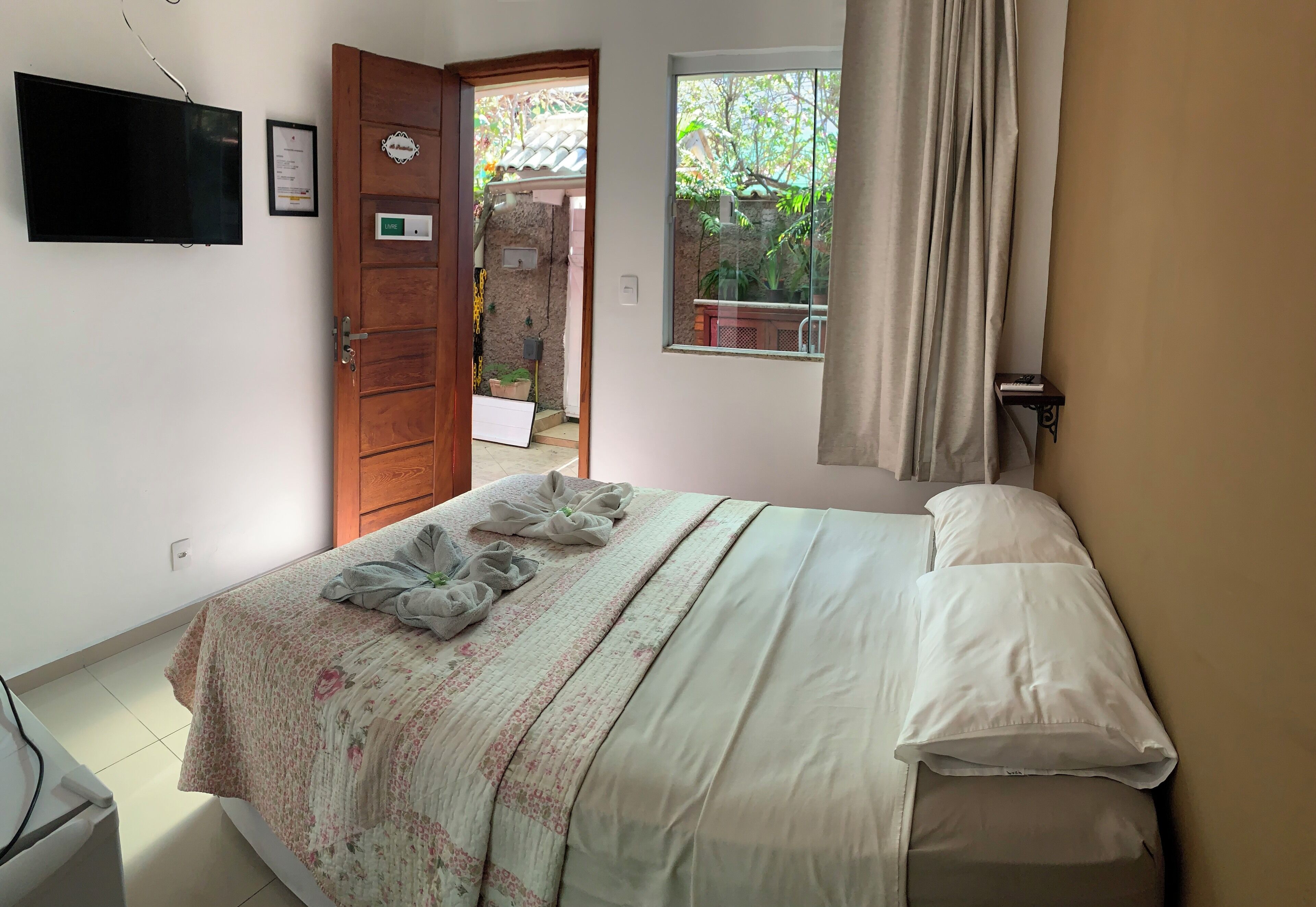 A bedroom with a bed, a TV mounted on the wall, a window with a view of greenery, and a door leading to an outdoor area.