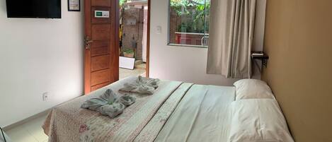 A bedroom with a bed, a TV mounted on the wall, a window with a view of greenery, and a door leading to an outdoor area.