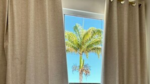 A neatly made bed with white linens and a brown blanket, a pair of white bathrobes on the bed, and beige curtains framing a window with a view of a palm tree. - Pousada Bougainville Suites (Arraial do Cabo)