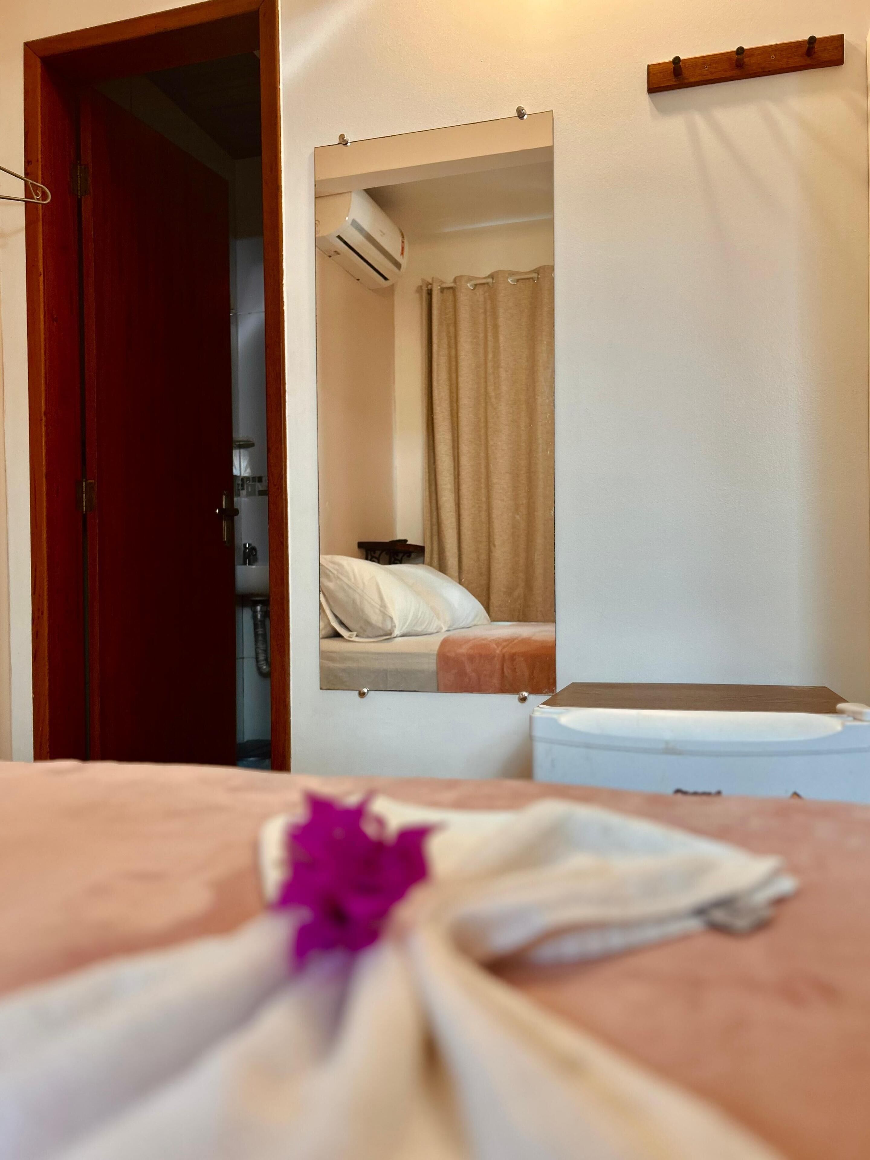 A hotel room with a bed, a small table with a flower, a mirror, and a wooden shelf.