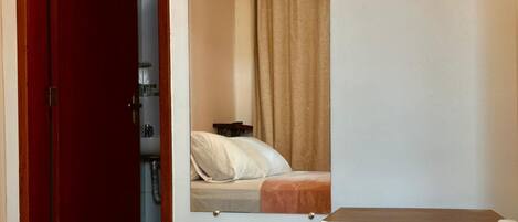 A hotel room with a bed, a small table with a flower, a mirror, and a wooden shelf.