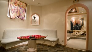Un espacio de spa con un banco acolchado blanco, una mesa de vidrio con flores, una ventana de arco montada en la pared y un cuadro visible a través de un arco.