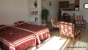 A room with two beds, a bookshelf, a dining table with chairs, and a kitchen area.