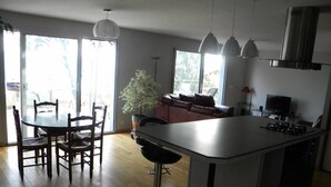 Une cuisine moderne avec un îlot central, des tabourets de bar, une table à manger avec des chaises, une plante en pot et un téléviseur.