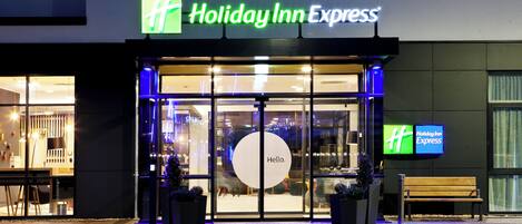 Exterior de un hotel Holiday Inn Express con un letrero iluminado grande, una entrada de vidrio y un banco afuera.