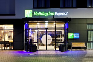 Exterior de un hotel Holiday Inn Express con un letrero iluminado grande, una entrada de vidrio y un banco afuera.