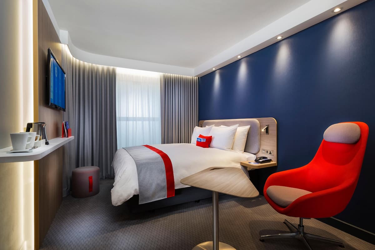 A modern hotel room with a bed, a red chair, a TV, and a small table with cups and a remote control.