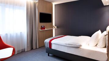 Una habitación de hotel moderna con una cama, un televisor montado en la pared, una silla roja y una ventana con cortinas.