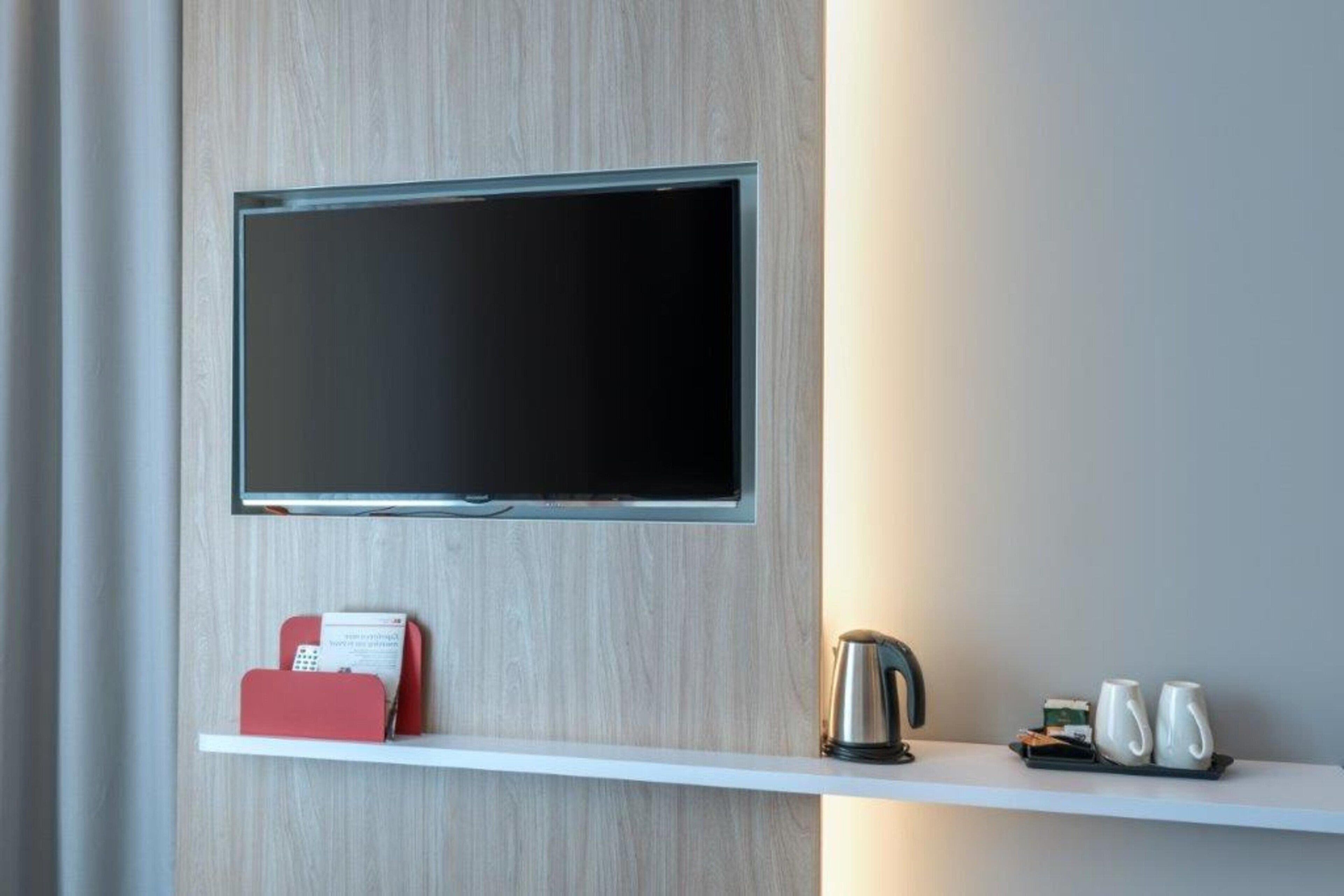 Un téléviseur à écran plat est fixé au mur, lequel est agrémenté d’un panneau en bois. Sous le téléviseur, on trouve une étagère blanche sur laquelle sont posés un téléphone rouge, une bouilloire argentée et trois tasses blanches.