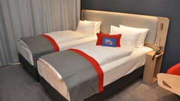 A hotel room with two beds, each with white bedding and red accents, a wooden nightstand with a phone, and a desk with an orange chair.