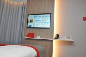A hotel room with a bed, a mounted TV displaying a menu, a shelf with a kettle and cups, and a red accent chair.