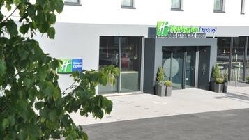 Een modern gebouw met een glazen gevel, een groen bord met de tekst 'Holiday Inn Express' en een uitnodigende ingang met een 'Hallo'-bord.