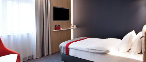 A hotel room with a bed, a TV mounted on the wall, a red chair, and a window with curtains.