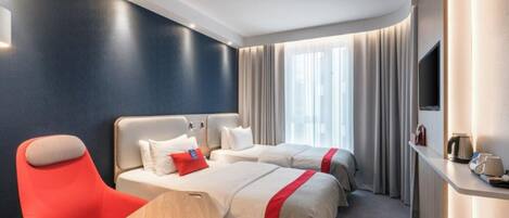 A hotel room with two beds, a red chair, a TV, and a small table with a kettle.