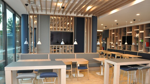 A modern restaurant with wooden tables and chairs, a large TV screen, and pendant lighting. - Holiday Inn Express Sindelfingen by IHG (Sindelfingen)
