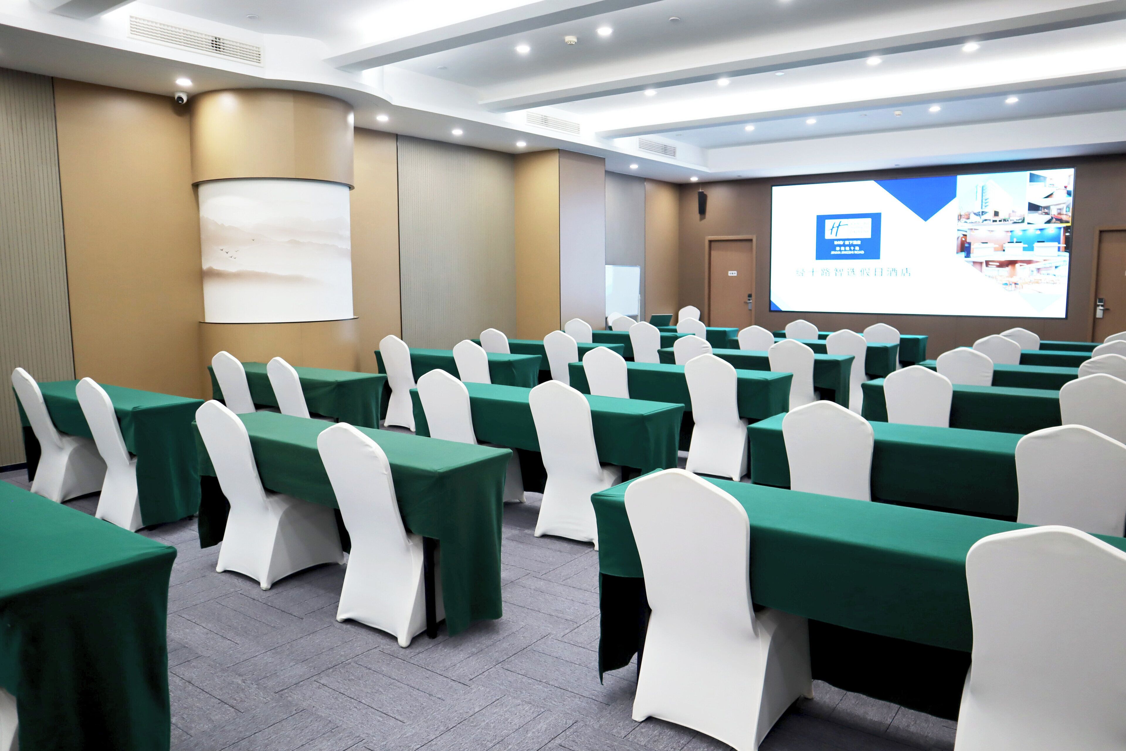 A conference room with rows of tables and chairs, a projector screen, and a presentation slide.
