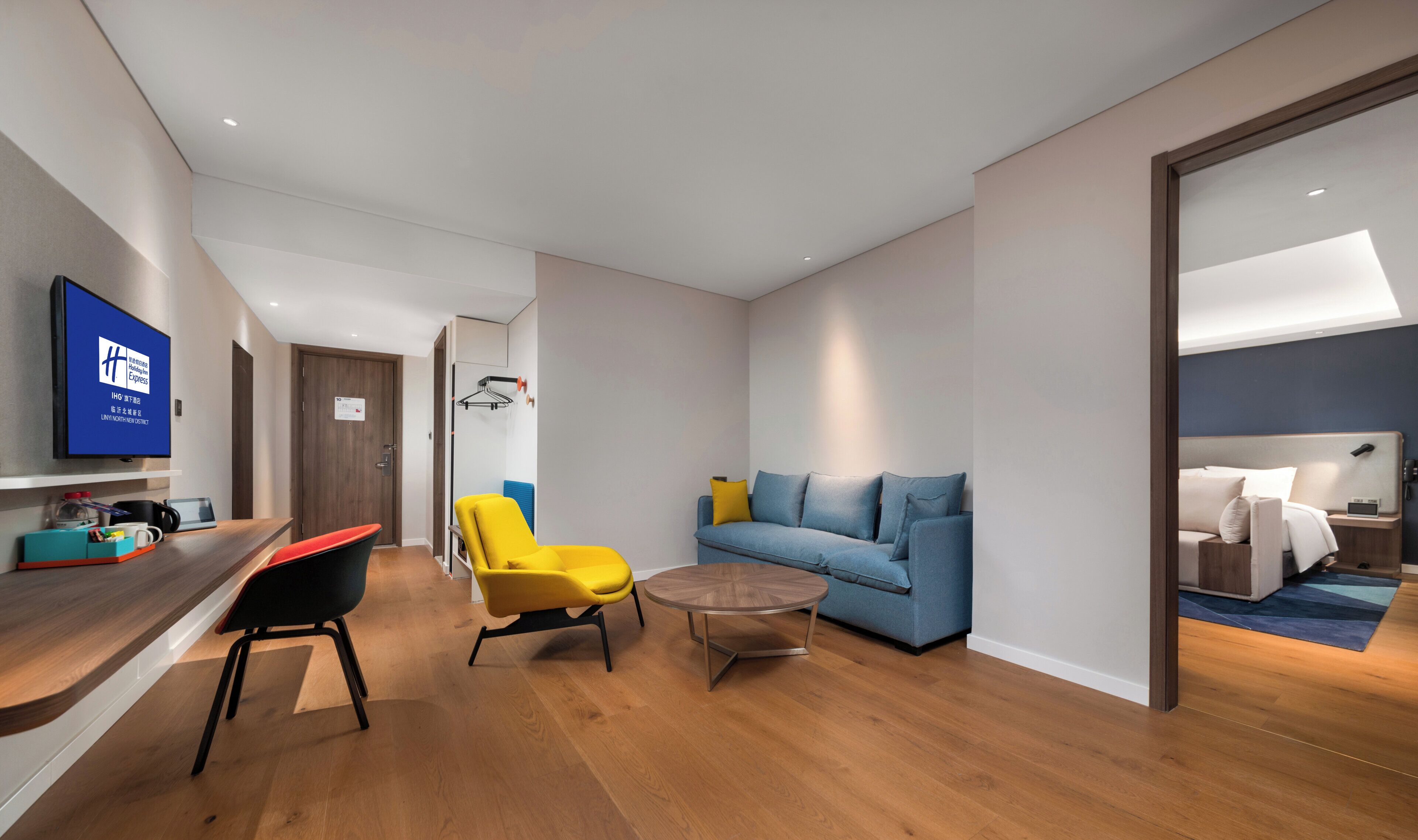 A modern hotel room with a flat-screen TV, a sofa, a yellow armchair, and a small round table.