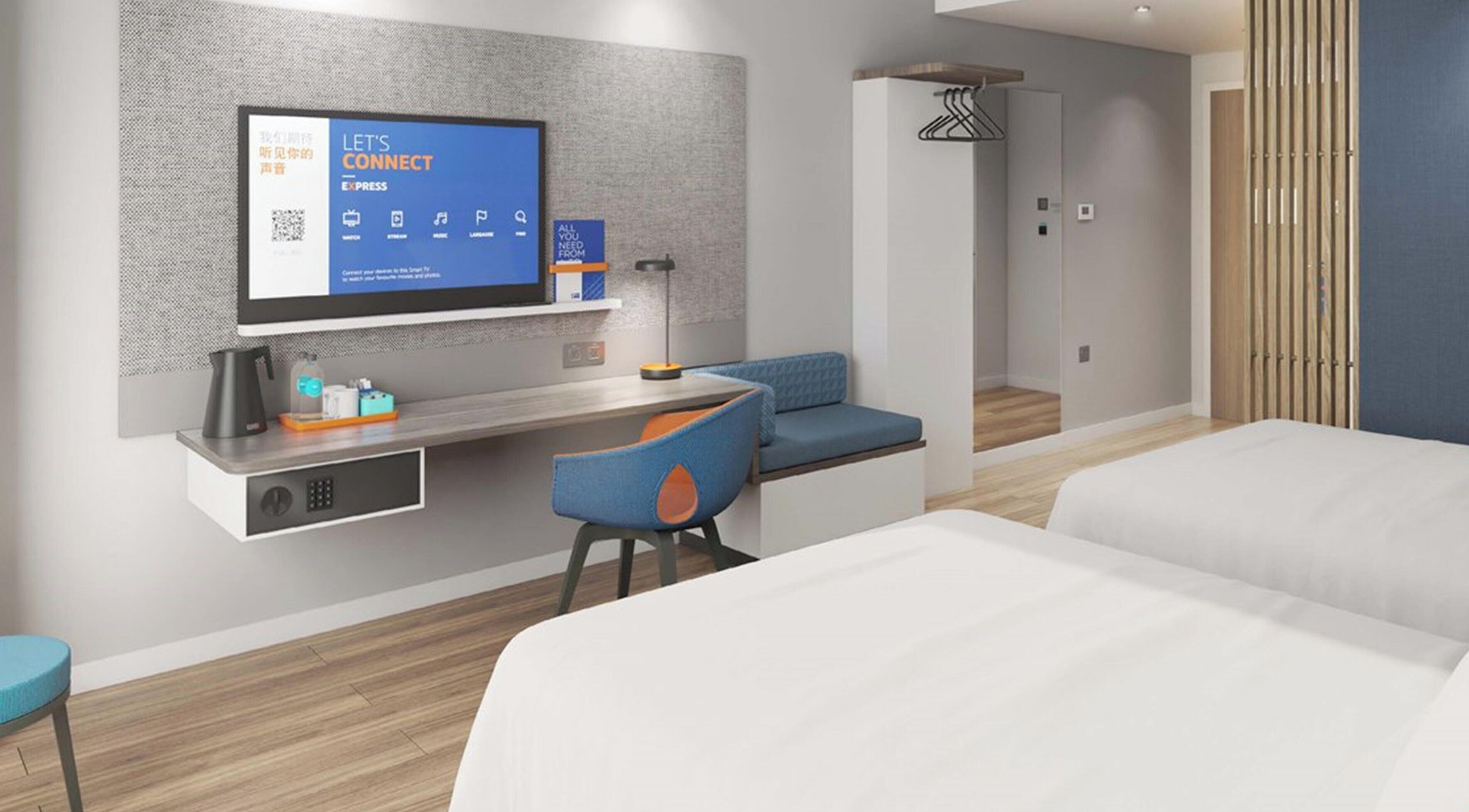 Hotellihuone, jossa on sänky, työpöytä tietokoneella, oleskelualue ja televisio, jossa näkyy QR-koodi.