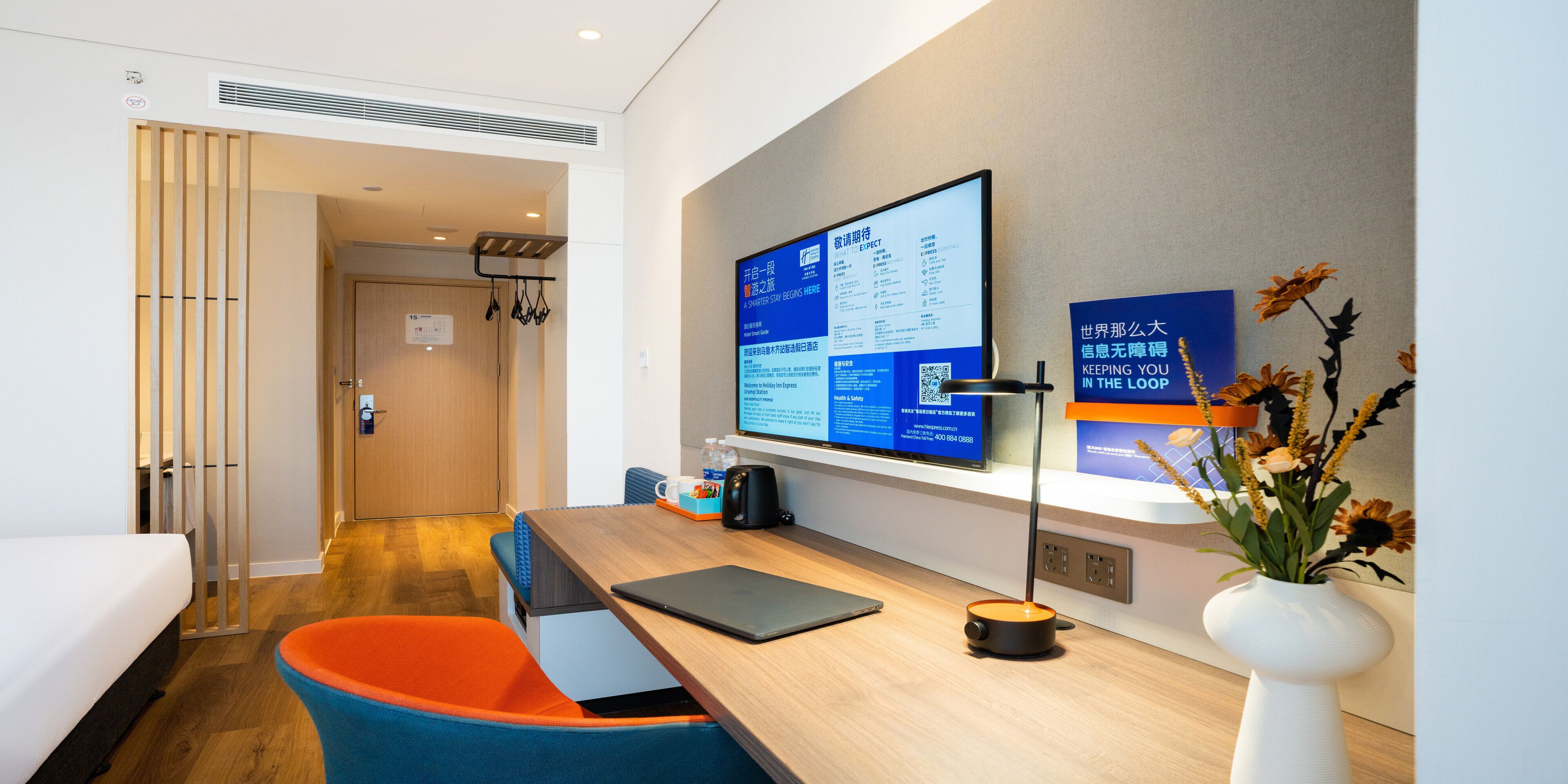 A modern hotel room with a wooden desk, a laptop, a vase with flowers, and a large monitor displaying information.