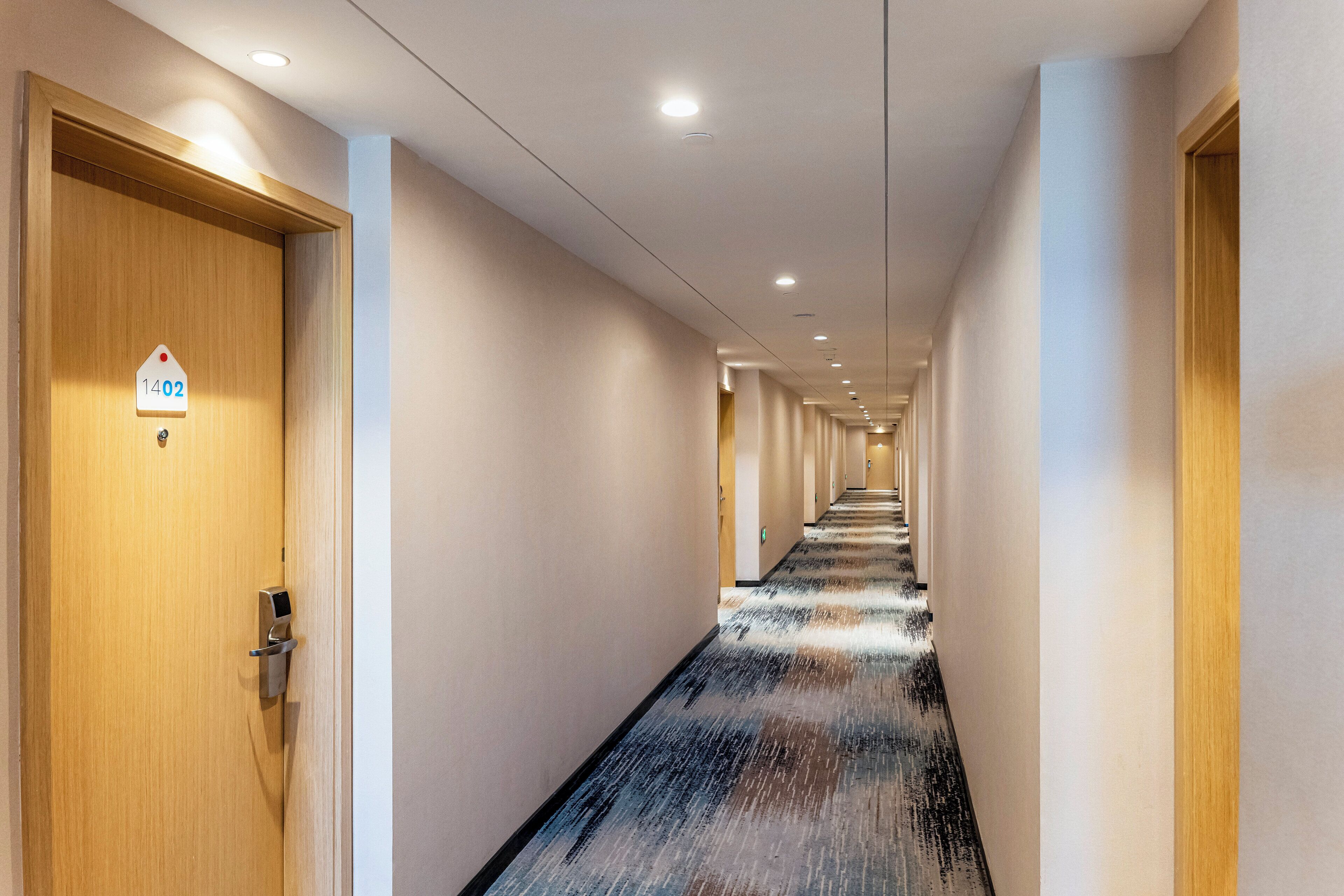 A hotel hallway with a wooden door on the left, a sign with the number 1402, and a long corridor with doors on both sides.