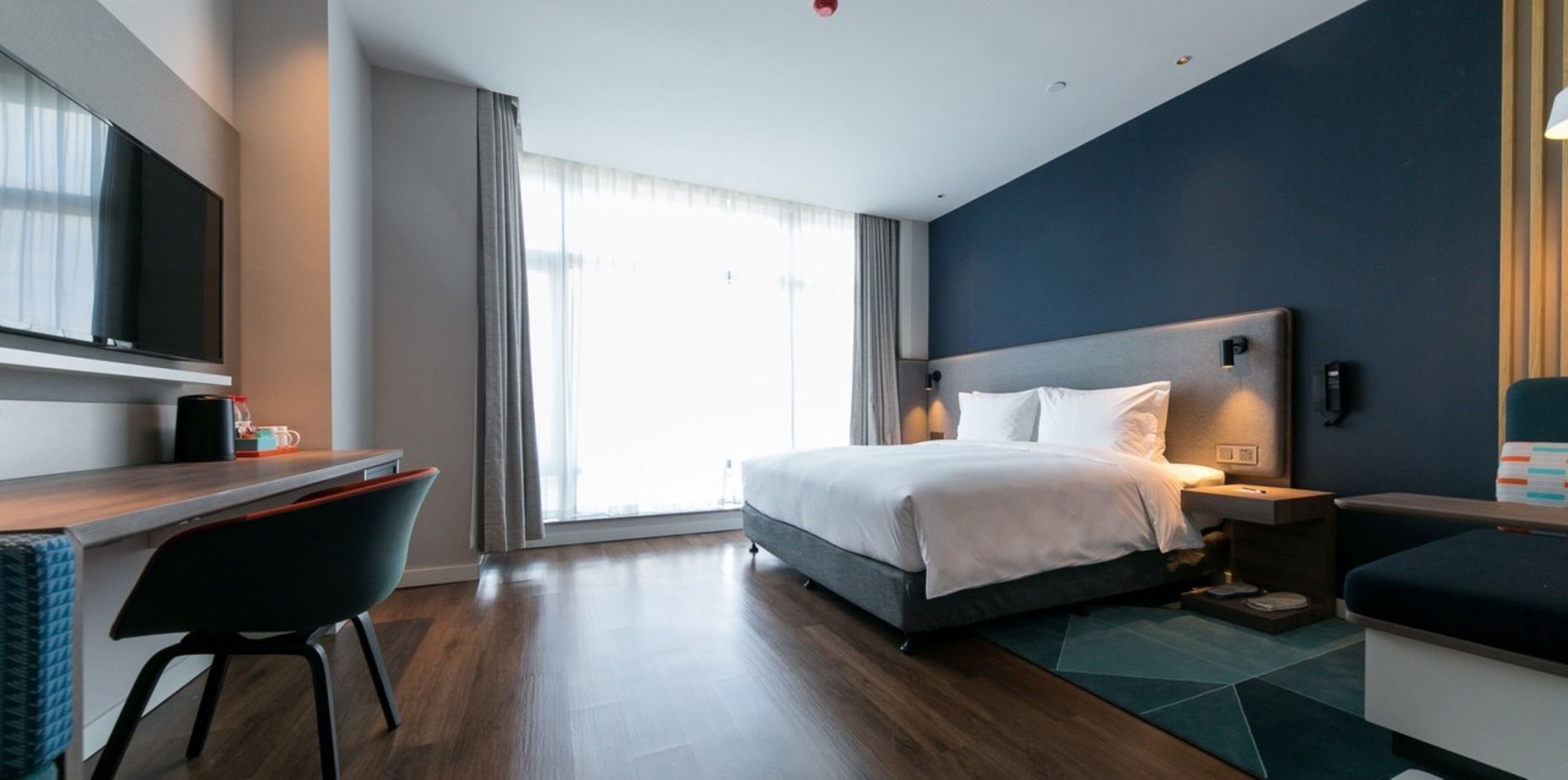 A modern hotel room with a bed, bedside table, desk, chair, and a large window.