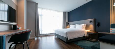 A modern hotel room with a bed, bedside table, desk, chair, and a large window.