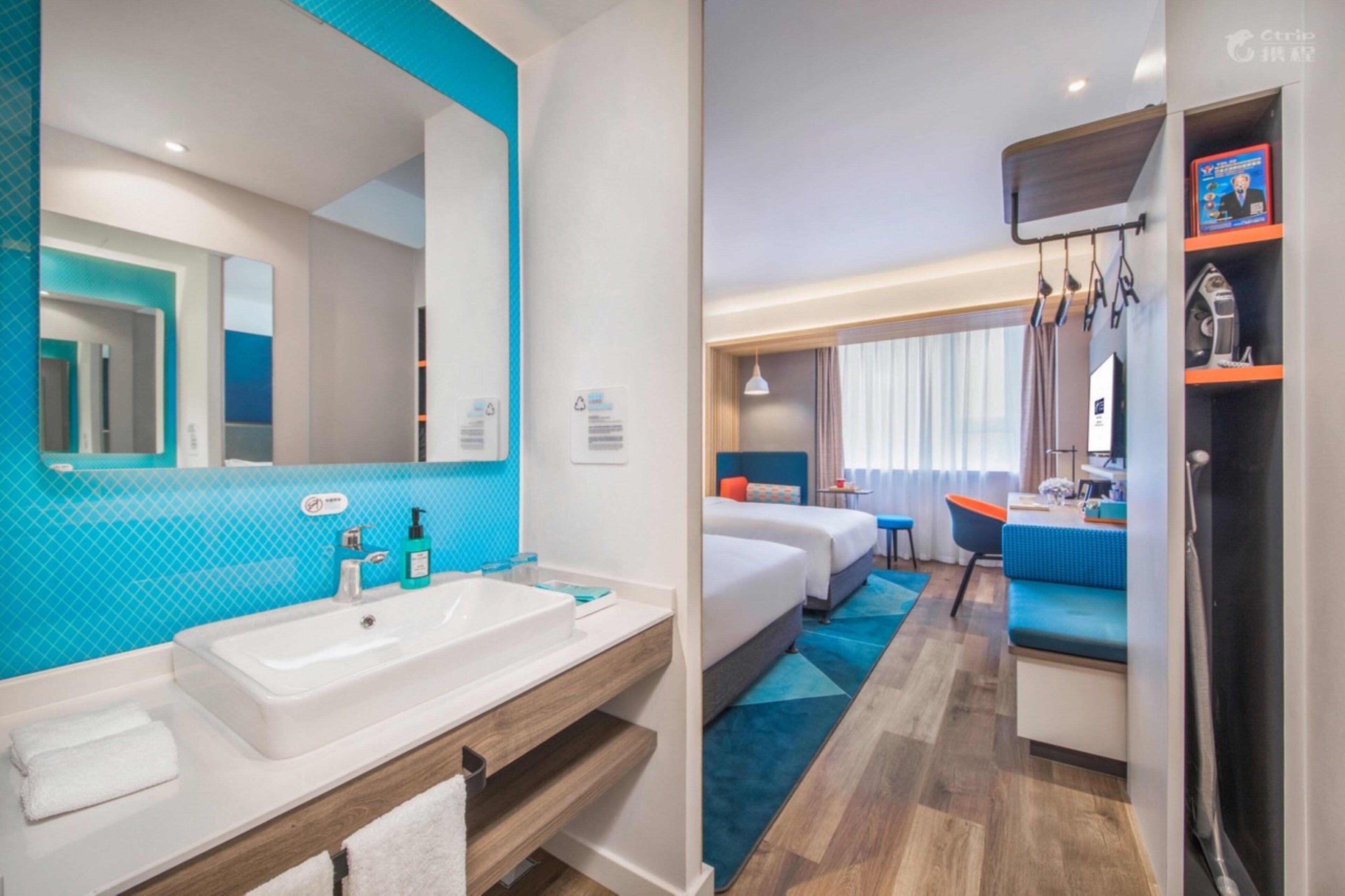Habitación de hotel con un espejo grande en el baño, un lavamanos blanco y una pared de acento azul. Hay dos camas con ropa de cama azul y un escritorio pequeño con una silla.