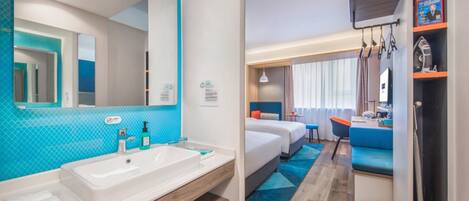 Habitación de hotel con un espejo grande en el baño, un lavamanos blanco y una pared de acento azul. Hay dos camas con ropa de cama azul y un escritorio pequeño con una silla.