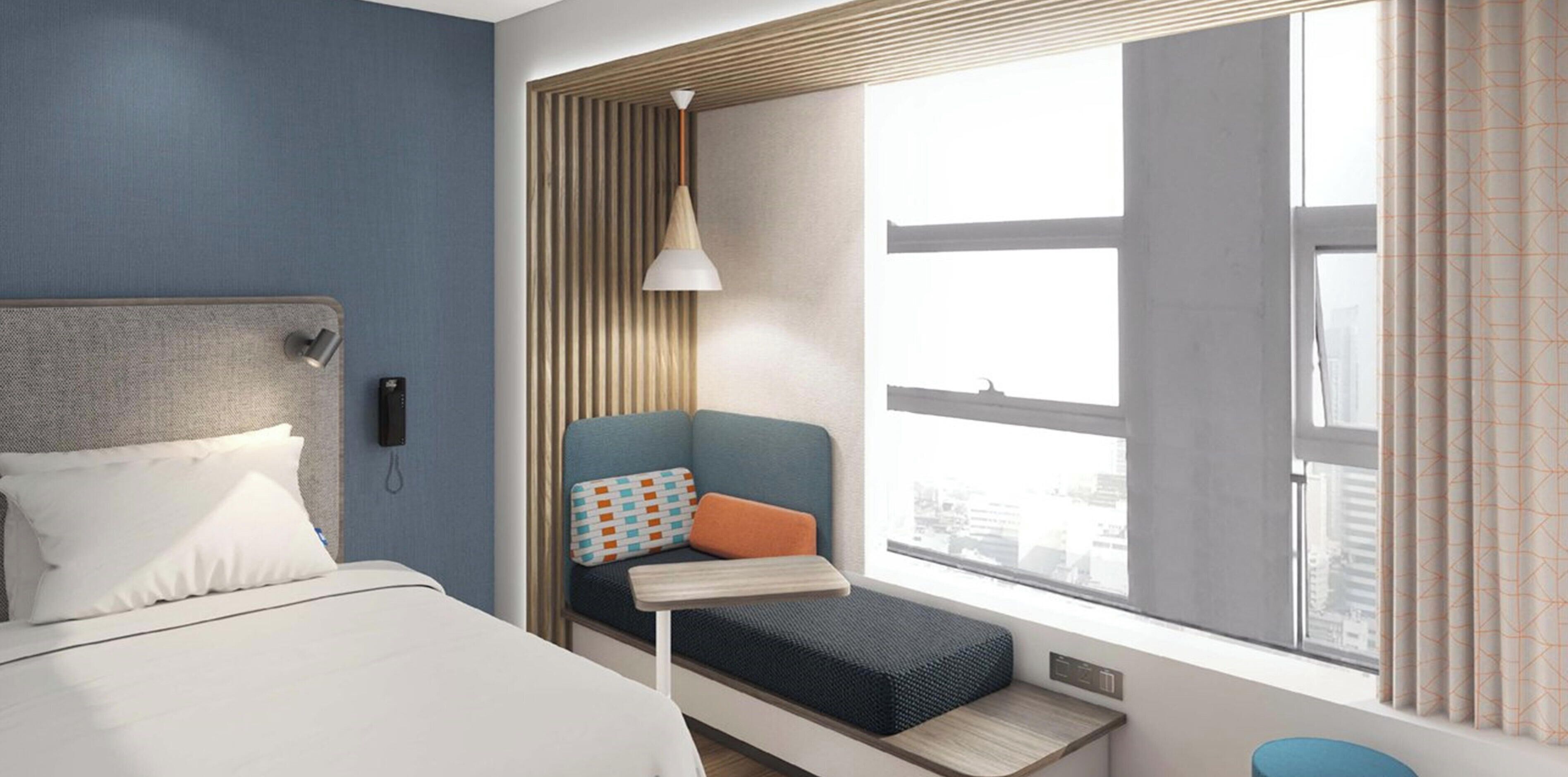 A hotel room with a bed, a small seating area with a pillow, a lamp, and a window with curtains.