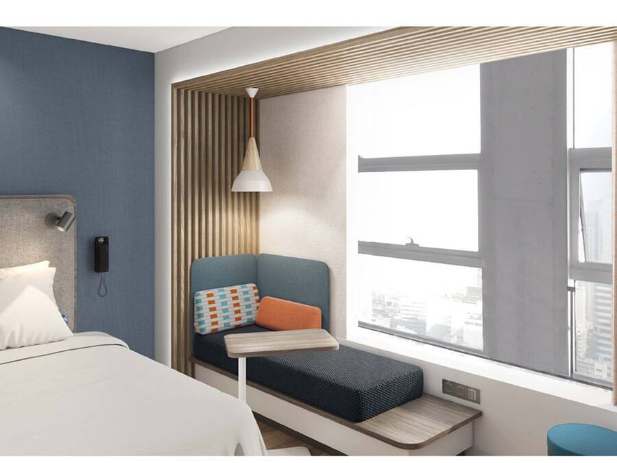 Una habitación de hotel moderna con una cama, un pequeño rincón de descanso con un banco y cojines, un escritorio y un amplio ventanal con cortinas.