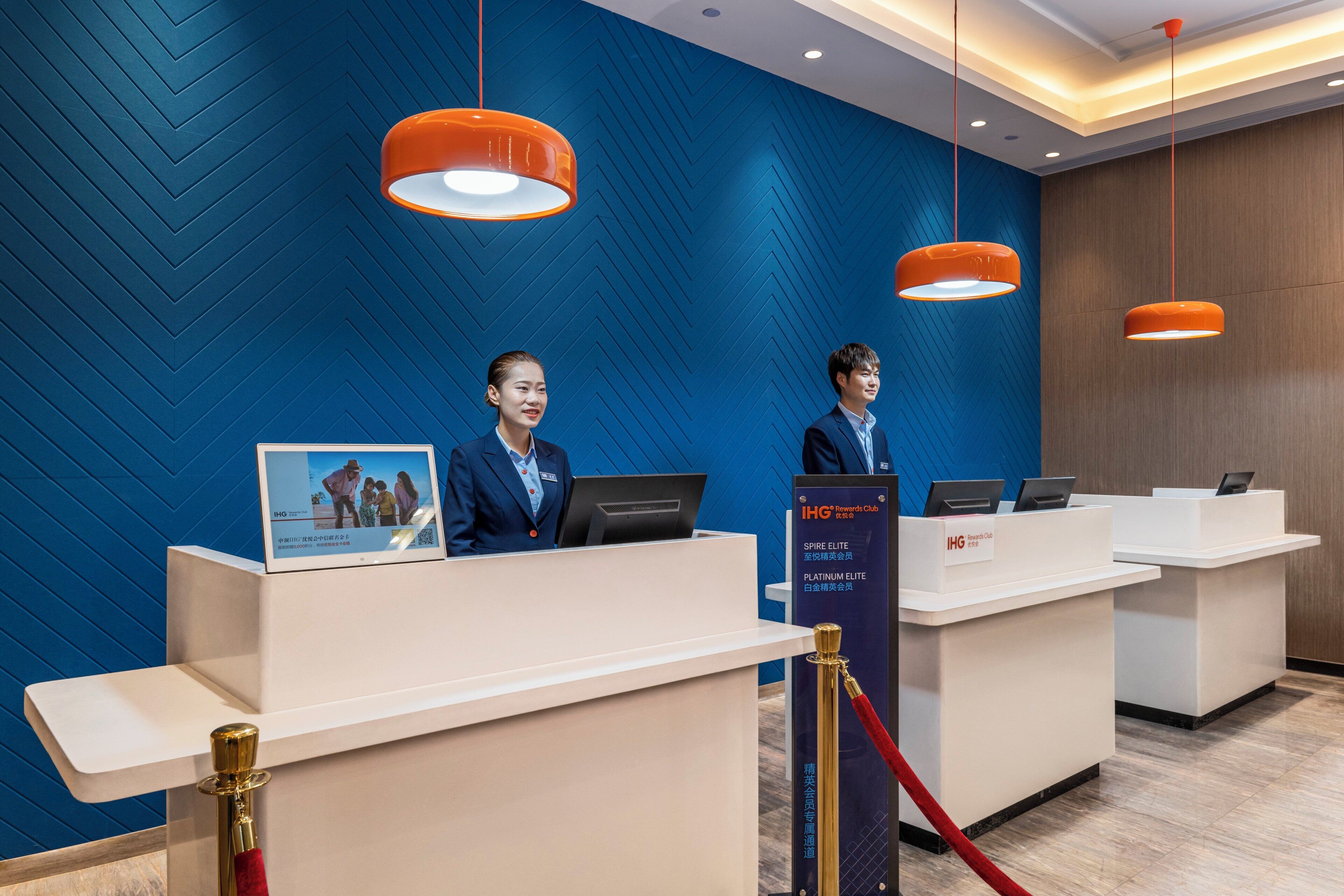 A hotel reception desk with two staff members, a blue patterned wall, and orange pendant lights.