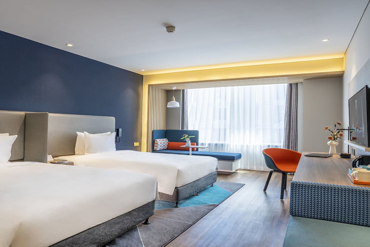 A hotel room with two beds, a desk with a chair, a television, and a window with curtains.
