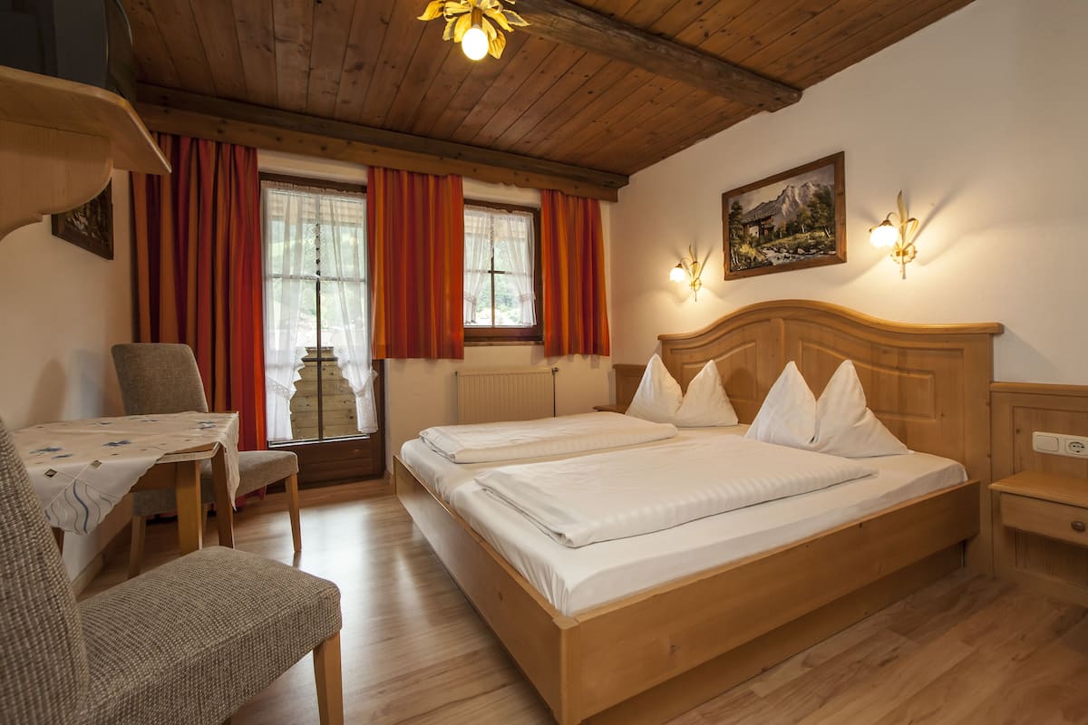 A wooden bed with white linens, a wooden desk, and a chair in a room with wooden floors and a sloped ceiling.