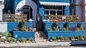 A two-story house with a blue exterior, a front porch, and a small garden with various plants. - Located in downtown O'side - Blue Jay by the Pier (Oceanside)